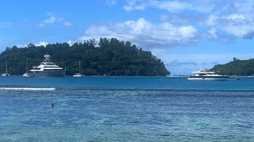 blue sea with two yachts anchored near a tree-covered island, clear sky above.