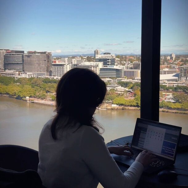 person sitting at a desk with a laptop, looking out over a river and city skyline through a large window.