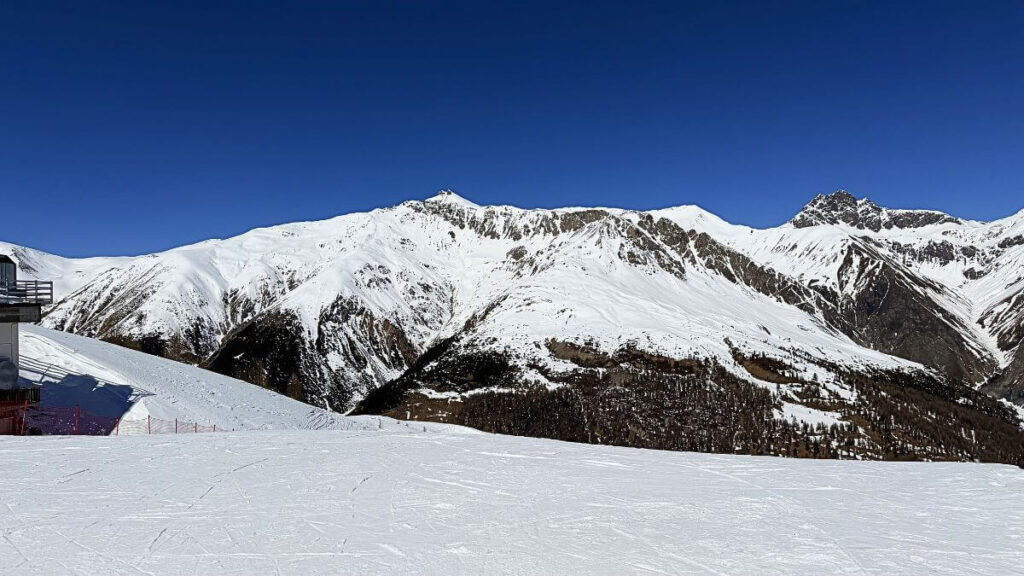 livigno-blue-bird-ski-day-empty-piste | Journey Beyond 40 snow-covered mountain range under a clear blue sky, with a ski slope and a small building on the left.