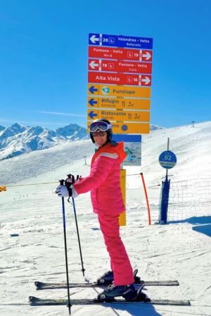 female skier in a pink suit with helmet and poles stands on a snowy slope beside a tall colorful signpost.